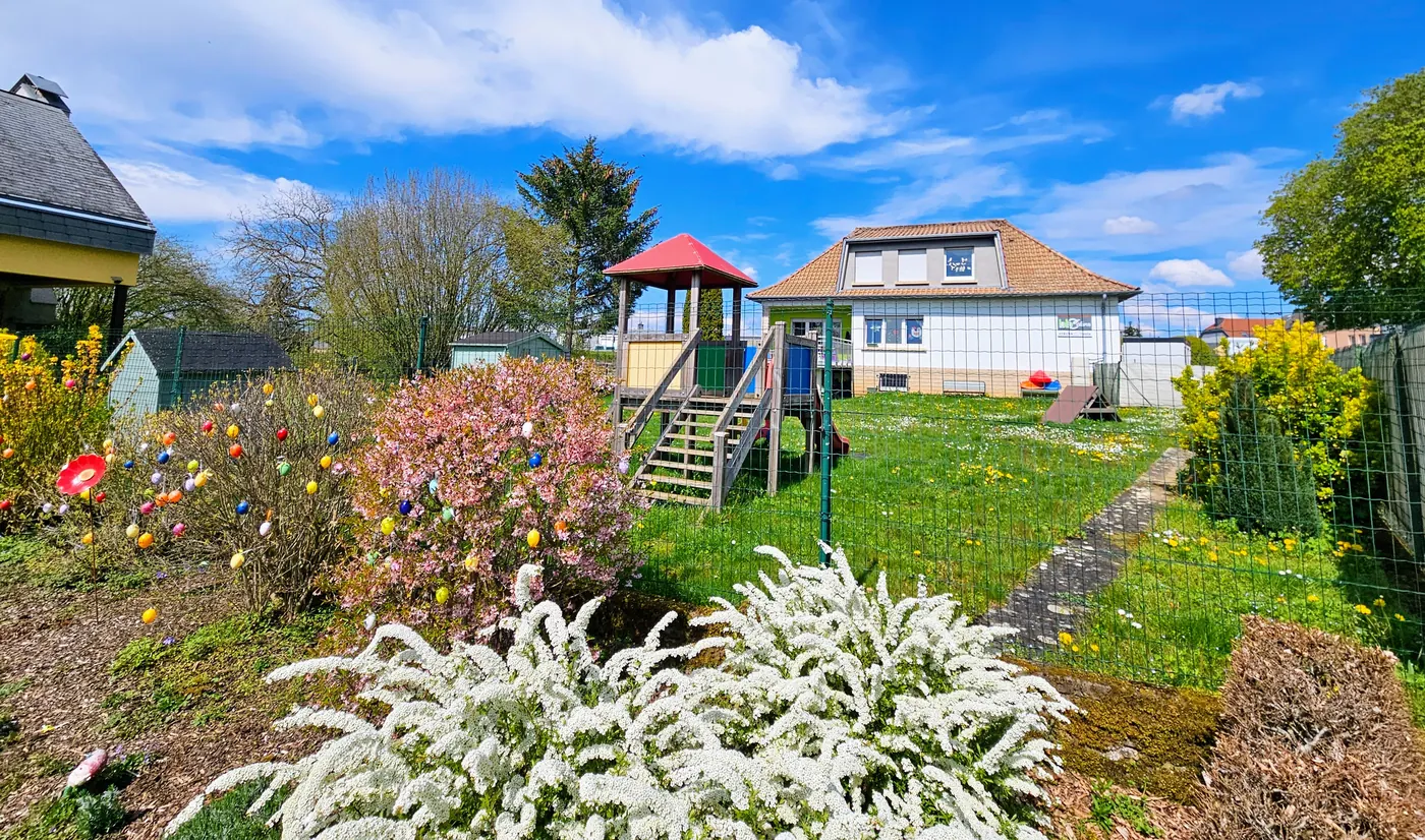 The nursery garden in spring, with flower beds, a playhouse and slide, and the house in the background